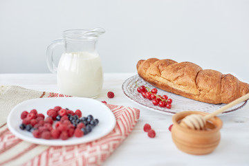 Different ingredients for breakfast. Croissants on plate on the wood board, berries, coffee, milk and honey on a white tablecloth. Traditional French breakfast. Top view