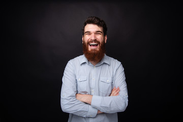Happiness and laughing. Portrait of a young bearded man standing with arms crossed on black background laughing