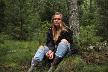 Gorgeous woman in outdoor outfit sits near tree in a scenic wood or forest full of green and feel relaxed