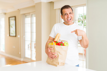 Middle age man holding groceries shopping bag at home very happy pointing with hand and finger