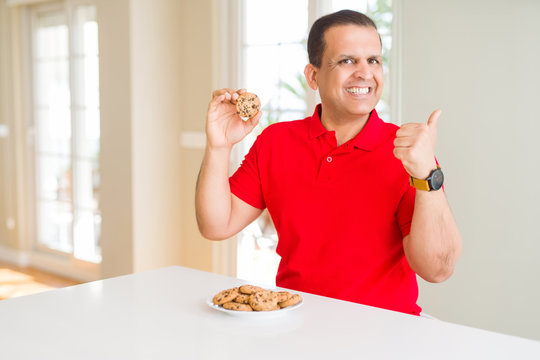 Middle age man eating chocolate chips cookies at home pointing and showing with thumb up to the side with happy face smiling