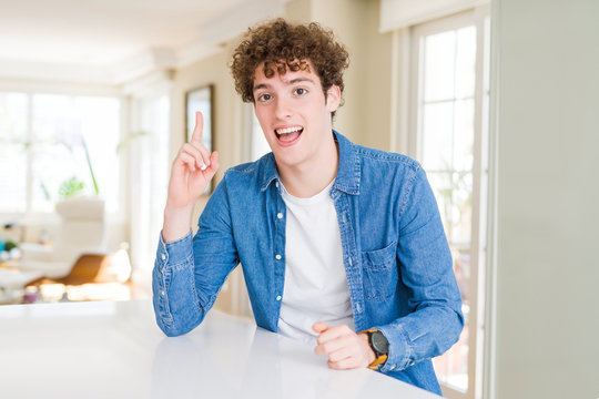 Young handsome man wearing casual denim jacket at home pointing finger up with successful idea. Exited and happy. Number one.