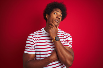 Fototapeta na wymiar Young african american man with afro hair wearing striped t-shirt over isolated red background with hand on chin thinking about question, pensive expression. Smiling with thoughtful face. 
