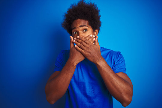 African american man with afro hair wearing t-shirt standing over isolated blue background shocked covering mouth with hands for mistake. Secret concept.