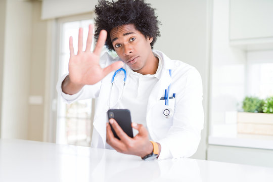 African American doctor man using smartphone at the clinic with open hand doing stop sign with serious and confident expression, defense gesture