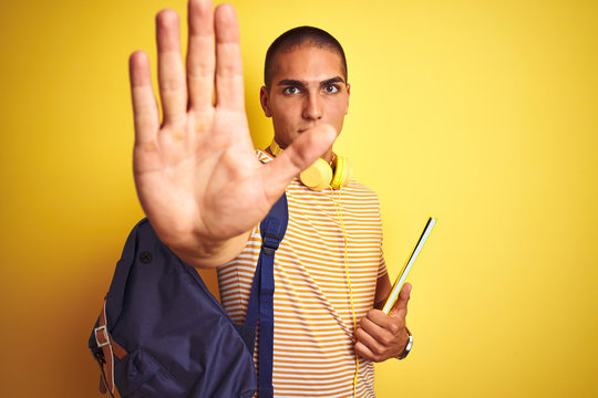 Young student man wearing headphones and backpack over yellow isolated background with open hand doing stop sign with serious and confident expression, defense gesture