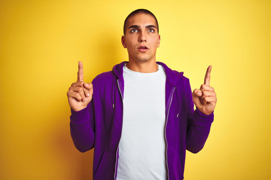 Young handsome man wearing purple sweatshirt over yellow isolated background amazed and surprised looking up and pointing with fingers and raised arms.