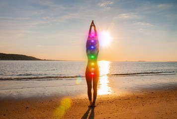 Woman is meditating with glowing seven chakras on the beach. Silhouette of woman is practicing yoga...