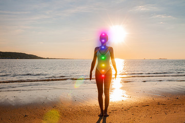 Woman is meditating with glowing seven chakras on the beach. Silhouette of woman is practicing yoga...