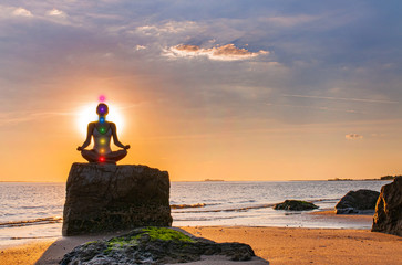 Woman is practicing yoga sitting on stone in Lotus pose at sunset. Silhouette of woman meditating...