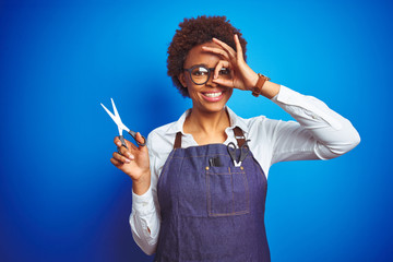 Obraz premium African american hairdresser woman holding scissors over blue isolated background with happy face smiling doing ok sign with hand on eye looking through fingers