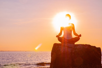 Woman is practicing yoga sitting on stone in Lotus pose at sunset. Silhouette of woman meditating...