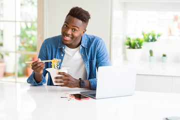 Obraz na płótnie Canvas Handsome young african business man eating delivery asian food and working using computer, enjoying noodles smiling