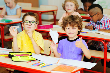 two little preschool boys sitting at a Desk or school table in the classroom holding paper...