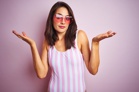 Beautiful woman wearing striped pink swimsuit and heart shaped sunglasses over pink background clueless and confused expression with arms and hands raised. Doubt concept.