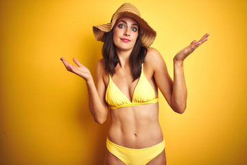 Beautiful woman wearing yellow bikini and summer hat over isolated yellow background clueless and confused expression with arms and hands raised. Doubt concept.