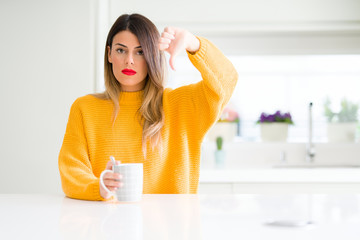 Young beautiful woman drinking a cup of coffee at home with angry face, negative sign showing dislike with thumbs down, rejection concept