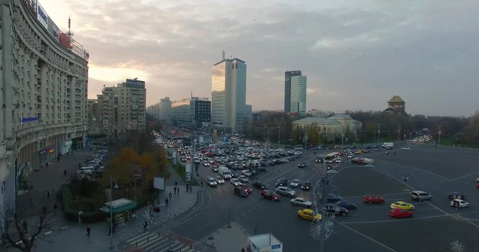 Aerial shot in Victoria square, one of the largest most busy traffic junctions in Bucharest. Drone lifts from the junction in the evening, golden sky, people crossing the street, cars shot passing by.