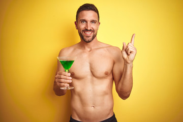 Fototapeta premium Young handsome shirtless man drinking a summer cocktail over isolated yellow background very happy pointing with hand and finger to the side