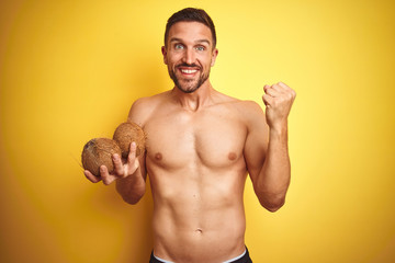 Fototapeta premium Young handsome shirtless man holding exotic tropical coconut over isolated yellow background screaming proud and celebrating victory and success very excited, cheering emotion
