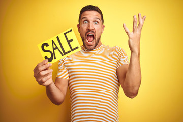 Young man holding sale advertising poster board over yellow isolated background very happy and excited, winner expression celebrating victory screaming with big smile and raised hands