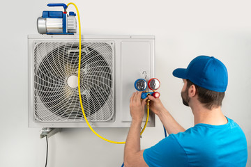 Installation service fix  repair maintenance of an air conditioner outdoor unit, by cryogenist technican worker evacuate the system with vacuum pump and manifold gauges in blue shirt baseball cap - obrazy, fototapety, plakaty