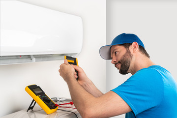 Installation service fix  repair maintenance of an air conditioner indoor unit, by cryogenist technican worker checking the temperature with infrared thermometer in blue shirt baseball cap