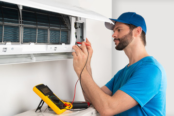 Installation service fix repair maintenance of an air conditioner indoor unit, by cryogenist technican worker with multimeter checking electric blue shirt baseball cap