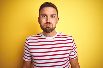 Young handsome man wearing casual red striped t-shirt over yellow isolated background puffing cheeks with funny face. Mouth inflated with air, crazy expression.