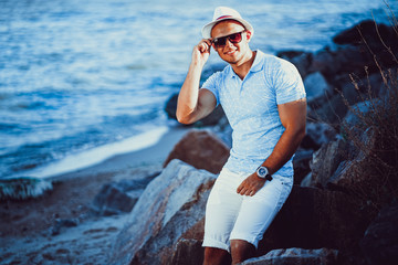 The man at the resort in a white shirt and sunglasses, hat sitting on a rock on the sea background.