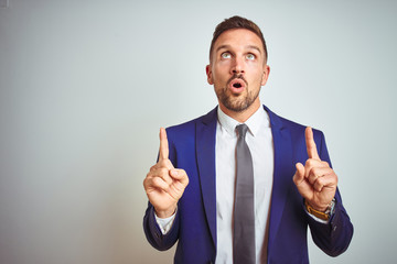 Young handsome business man over white isolated background amazed and surprised looking up and pointing with fingers and raised arms.