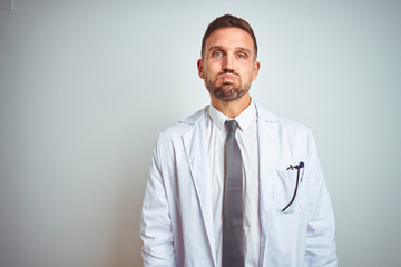 Young handsome doctor man wearing white profressional coat over isolated background puffing cheeks with funny face. Mouth inflated with air, crazy expression.
