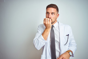 Young handsome doctor man wearing white profressional coat over isolated background looking stressed and nervous with hands on mouth biting nails. Anxiety problem.