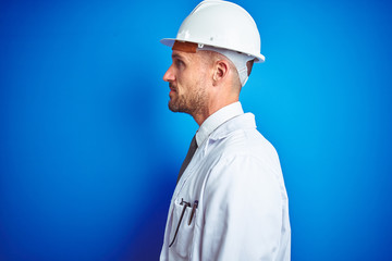 Young handsome engineer man wearing safety helmet over blue isolated background looking to side, relax profile pose with natural face with confident smile.
