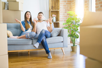 Young beautiful couple relaxing sitting on the sofa around boxes from moving to new house relax and smiling with eyes closed doing meditation gesture with fingers. Yoga concept.