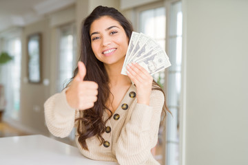 Young woman holding 20 dollars bank notes happy with big smile doing ok sign, thumb up with fingers, excellent sign