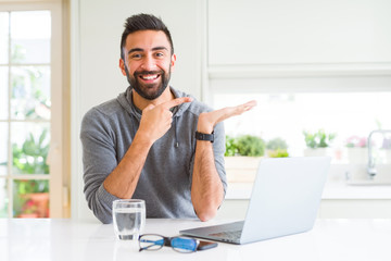 Handsome hispanic man working using computer laptop amazed and smiling to the camera while presenting with hand and pointing with finger.