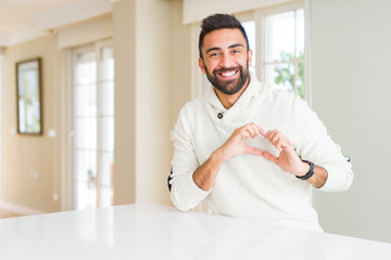 Obraz premium Handsome hispanic man wearing casual white sweater at home smiling in love showing heart symbol and shape with hands. Romantic concept.