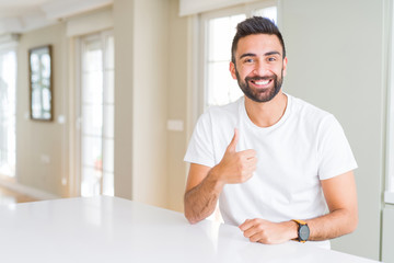 Handsome hispanic man casual white t-shirt at home doing happy thumbs up gesture with hand. Approving expression looking at the camera showing success.