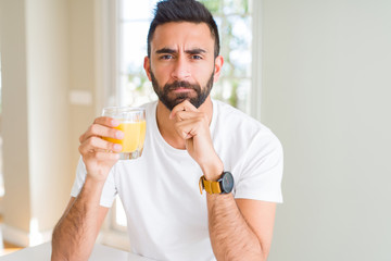 Handsome hispanic man drinking healthy orange juice serious face thinking about question, very confused idea