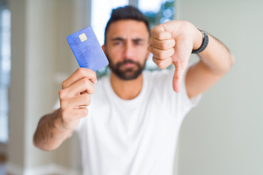 Handsome hispanic man holding credit card with angry face, negative sign showing dislike with thumbs down, rejection concept