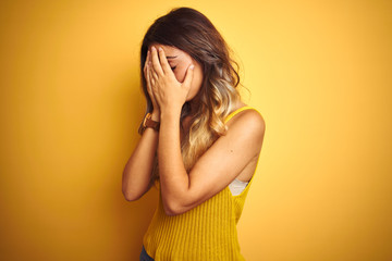 Young beautiful woman wearing t-shirt over yellow isolated background with sad expression covering...