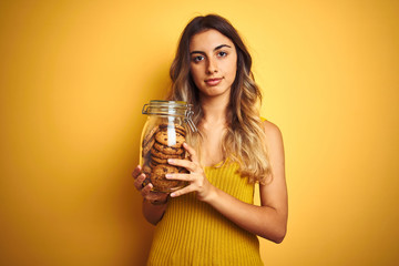 Young beautiful woman holding jar of cookies over yellow isolated background with a confident expression on smart face thinking serious
