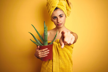 Young beautiful woman wearing a towel holding cactus pot over yellow isolated background pointing with finger to the camera and to you, hand sign, positive and confident gesture from the front