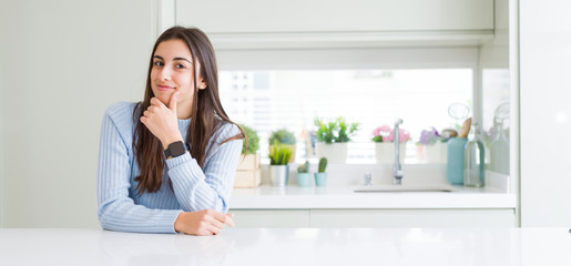 Wide angle picture of beautiful young woman sitting on white table at home looking confident at the camera with smile with crossed arms and hand raised on chin. Thinking positive.