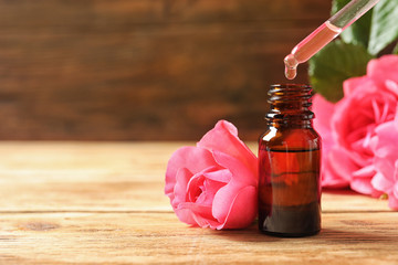 Bottle of rose essential oil, pipette and flowers on wooden table, space for text