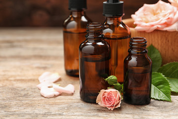 Bottles of rose essential oil and flowers on wooden table, space for text