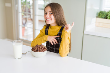 Beautiful young girl kid eating chocolate cereals and glass of milk for breakfast with a big smile...