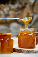 A table with a wicker table top, honey in honeycombs on a wooden board, honey flows from a spoon into an open glass jar. Medicinal herbs oregano, chamomile and strawberry flowers.