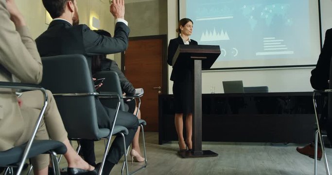 Caucasian female boss of the company standing at the lecturn in front of her colleagues and workers and having her annual speech or report with a video presentation of charts behind on the screen.